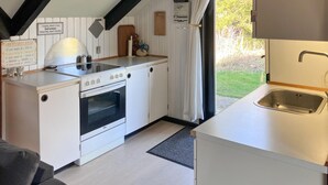 A compact kitchen with a stove, oven, and sink. There are signs on the wall, a window with curtains, and a view of greenery outside.