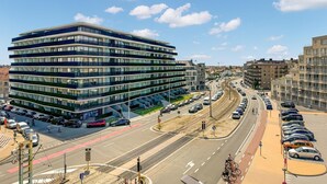 A modern multi-story building with a glass facade, located at a busy intersection with a tram line and a parking area. - Awesome apartment in Oostende (Oostende)