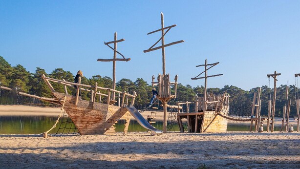 Een houten scheepsconstructie met een glijbaan, omringd door zand en water.