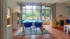 Een eethoek met een glazen tafel en blauwe stoelen, een gele fauteuil, en uitzicht op groen.
