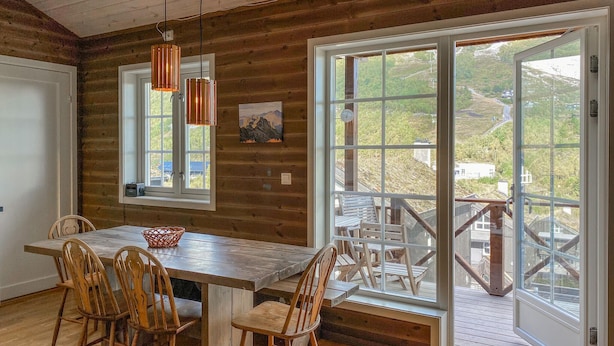 A wooden interior with a dining table, chairs, pendant lights, and a large window with a view of a hillside.