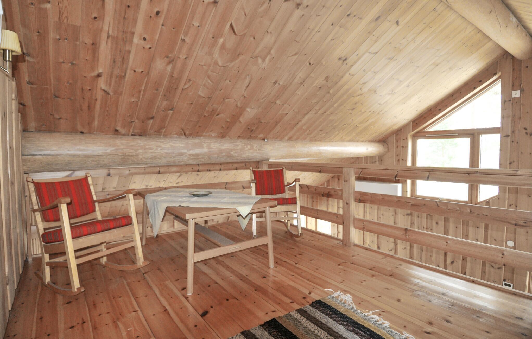 Eine Holz-Blockhütte mit Schlafzimmer im oberen Bereich, einem Holzboden, einem kleinen Tisch mit einer Schüssel, zwei Schaukelstühlen und Blick durch ein Fenster.