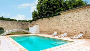 Piscina con muro de piedra y un puente en arco de metal. Hay tres sillas reclinables blancas.