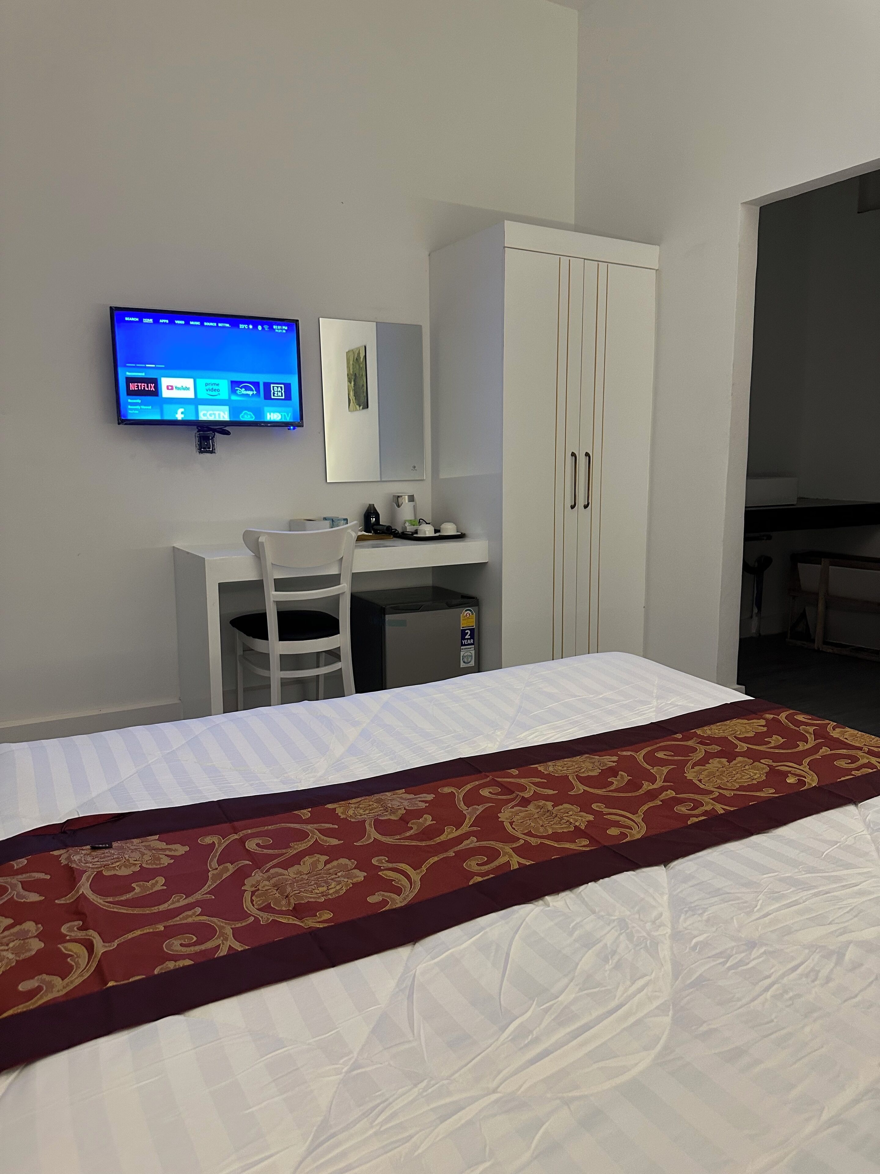 A hotel room with a bed, a desk with a chair, a television mounted on the wall, and a wardrobe.