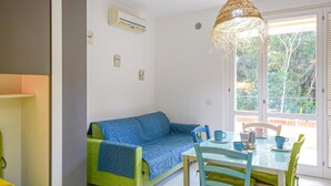 A cozy room with a green and blue sofa, a dining table with chairs, a hanging light fixture, and a window with a view of greenery. - Awesome apartment in Scarlino with WiFi (Scarlino)