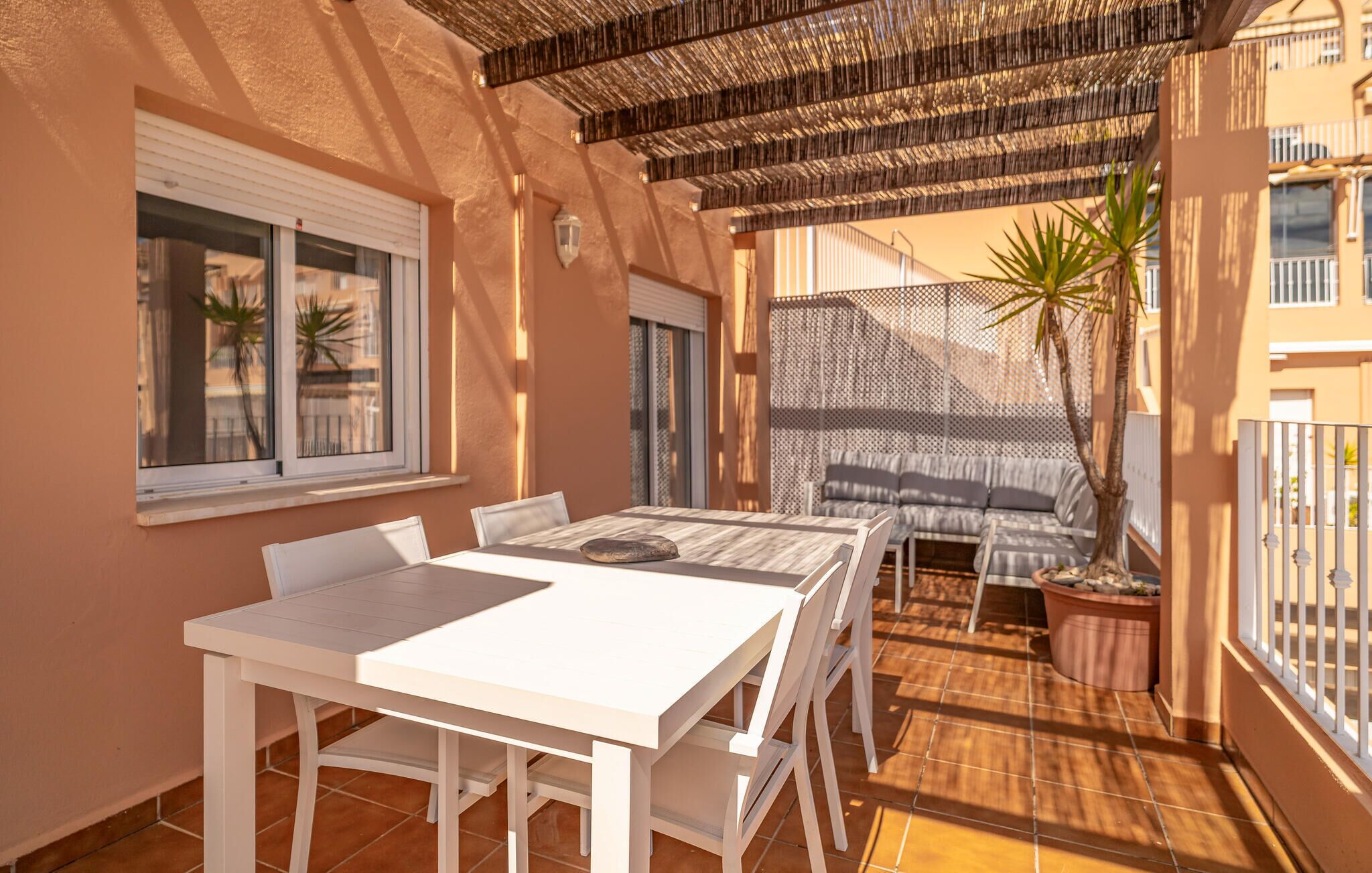 En solig uteplats med ett vitt bord och stolar, en mönstrad soffa och en pergola ovanför.