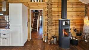 L’intérieur d’une cabane en rondins est agrémenté d’un poêle à bois central, d’une cuisine blanche aux meubles en bois et d’un escalier en bois menant à un étage supérieur.