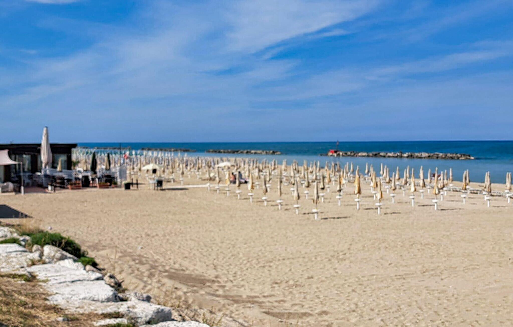 A beach with numerous umbrellas and lounge chairs, a building with outdoor seating, and a rocky shoreline.