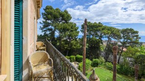 Ein Balkon mit Korbstuhl, Blick auf das Meer und ein Garten mit Bäumen und einer Lampe.