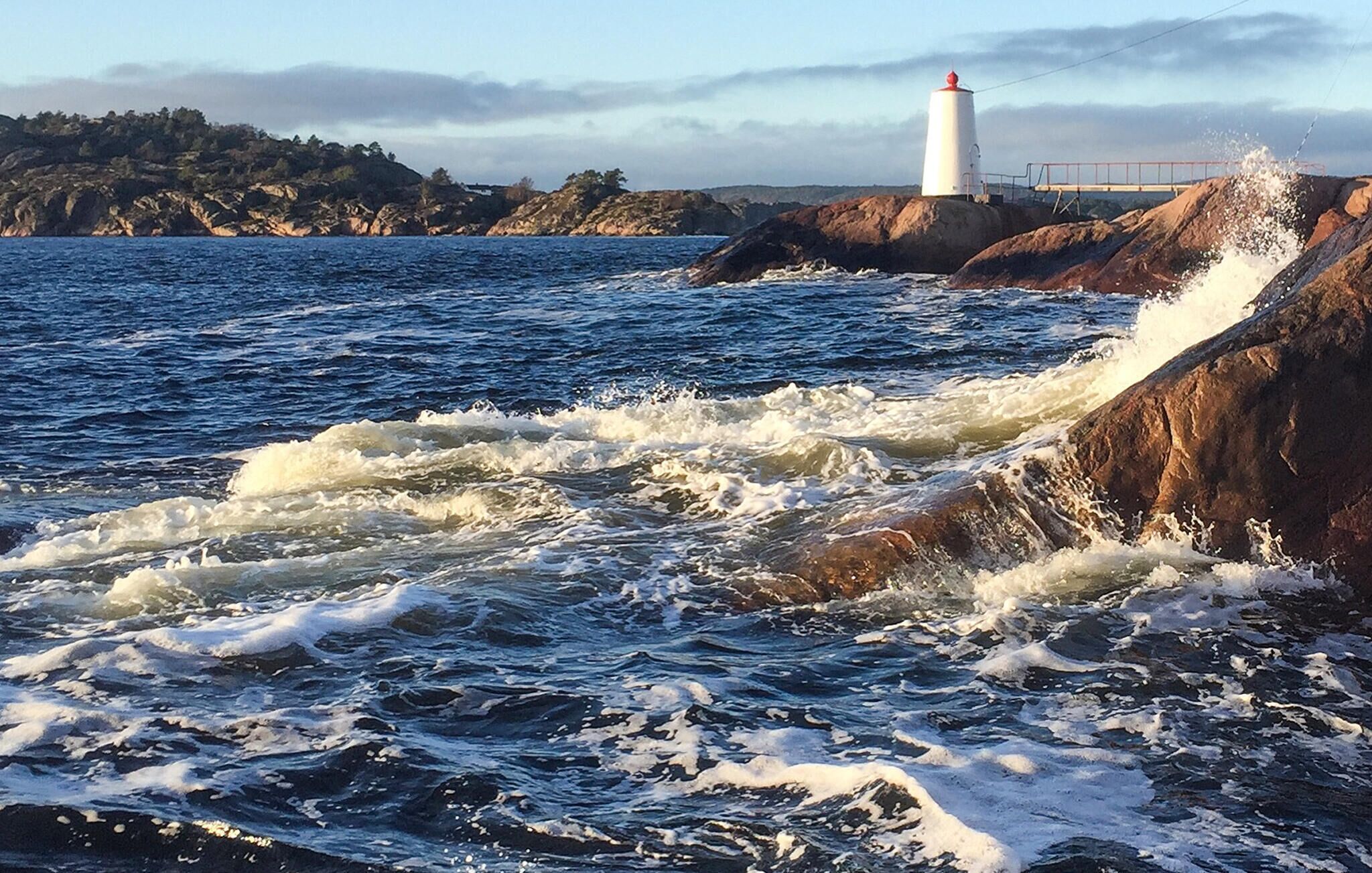 Une côte rocheuse où les vagues s’écrasent sur le rivage, avec un phare en arrière-plan.