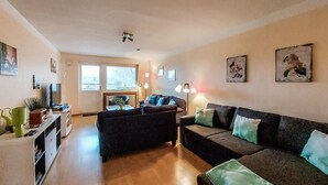 Un salón con un conjunto de sofá, una mesa de centro y un televisor. Hay dos ventanas con persianas, un ventilador de techo y lámparas de pared.