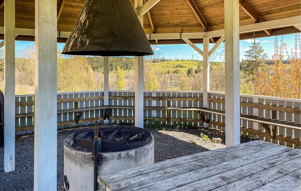 A covered outdoor area with a fire pit, wooden deck, and a view of a forested landscape.