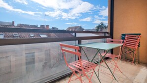 Ein Balkon mit Tisch und zwei Stühlen, der auf einen Stadtblick mit Gebäuden und blauem Himmel blickt.