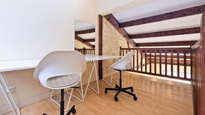 Ein moderner Arbeitsraum mit weißem Schreibtisch, zwei Stühlen und einem großen Spiegel. Das Zimmer zeichnet sich durch freiliegende Holzbalken und eine Steinwand aus.