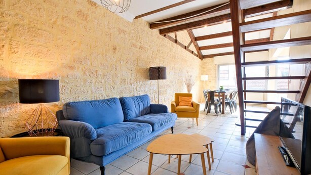 Ein gemütliches Wohnzimmer mit einer Steinwand, einem blauen Sofa, einem gelben Sessel, einem kleinen Holztisch und einem Essbereich mit Stühlen und Tisch.