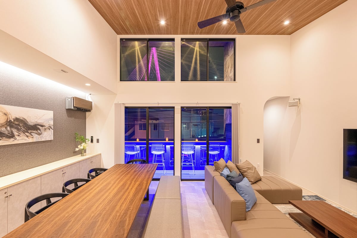 A modern living room with a wooden dining table, a sofa with cushions, and a TV mounted on the wall.