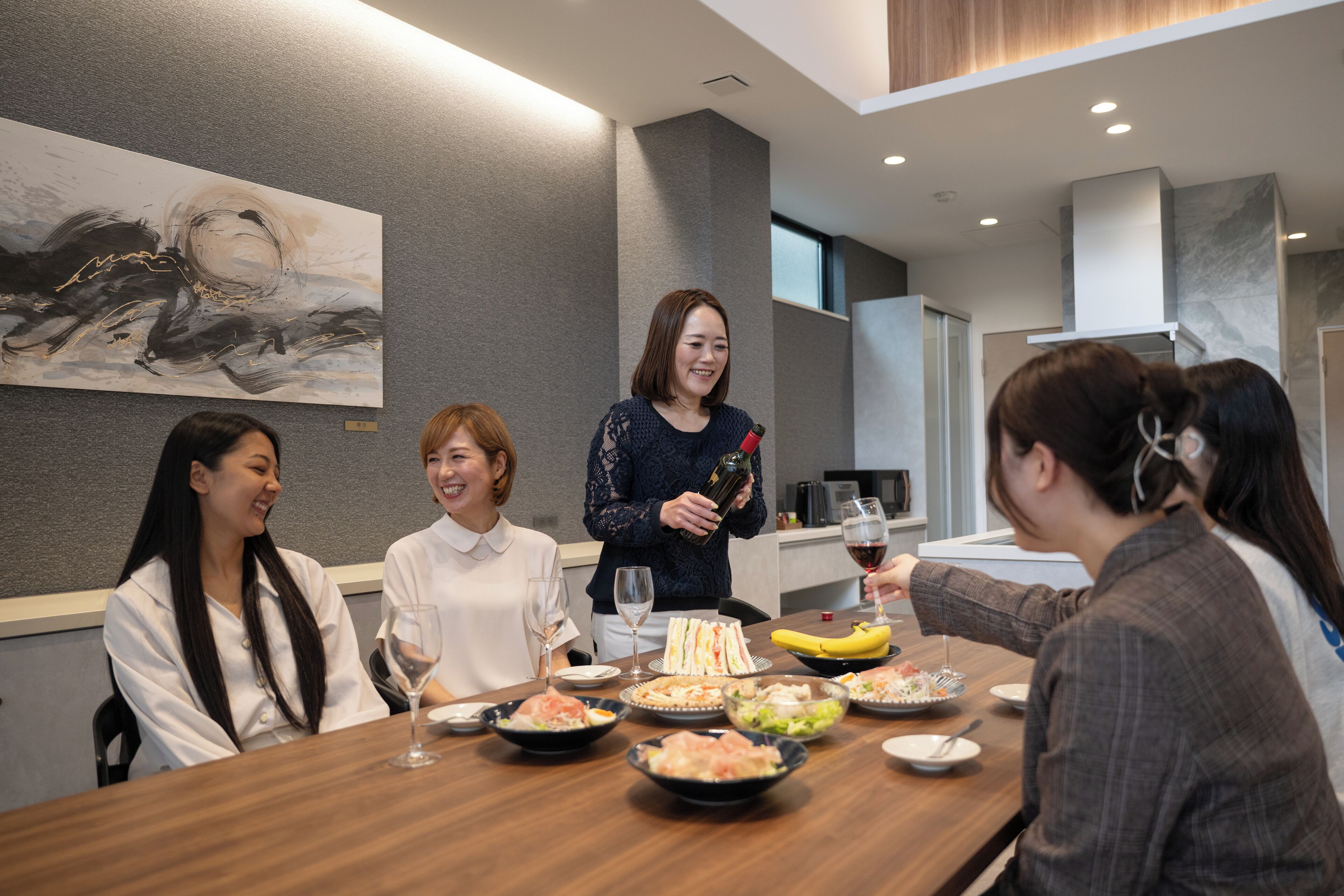 A group of people seated around a wooden table with various dishes and drinks, in a modern interior with a gray wall and a painting.