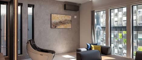 A spacious living area with a modern sofa set, a massage chair, and large windows offering a view of the city.