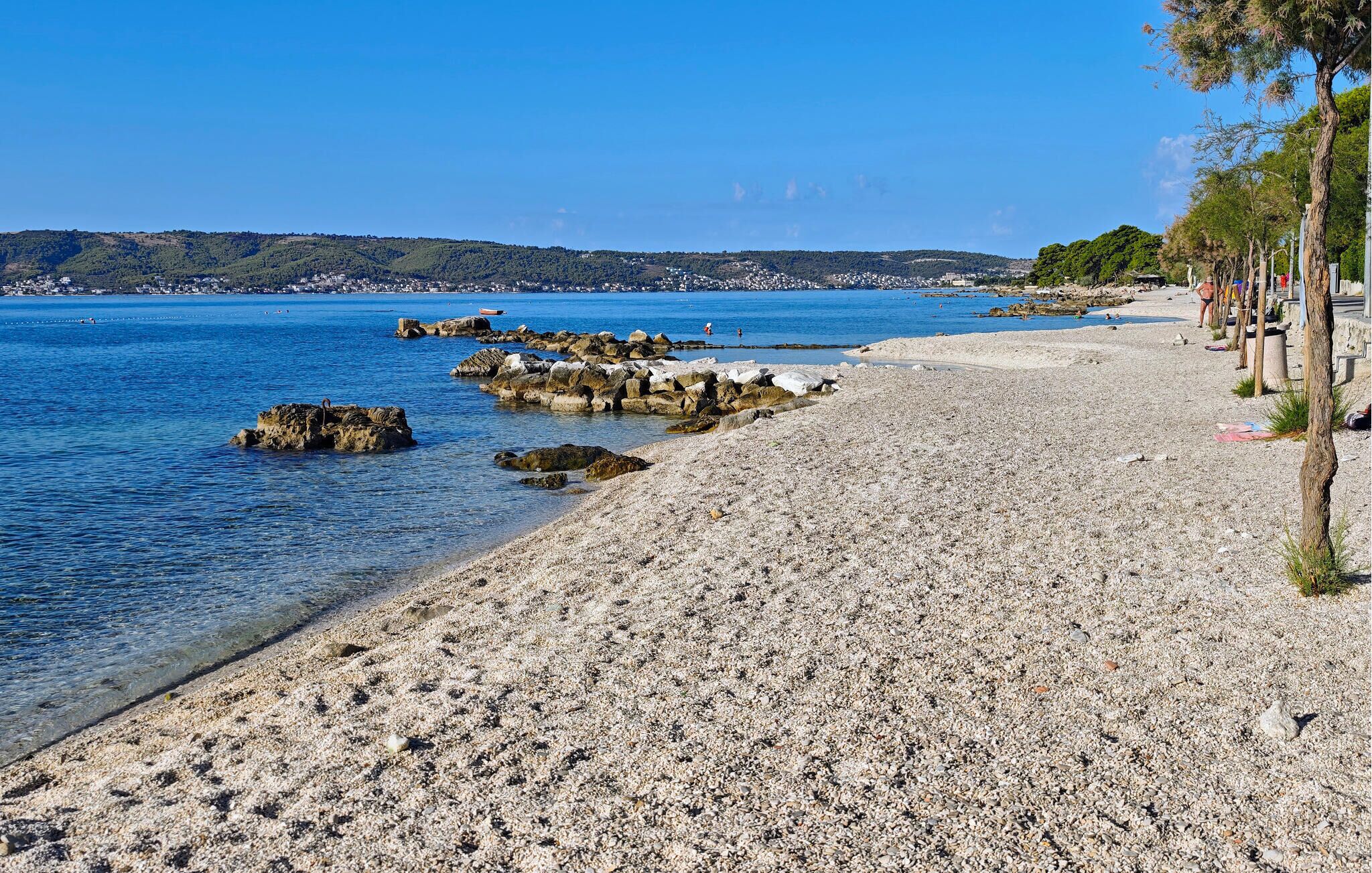 Une plage de galets bordée d’une eau d’un bleu limpide, avec des affleurements rocheux et une ville qui se devine au loin.