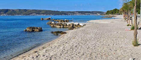 A pebble beach with clear blue water, rocky outcrops, and a distant town.