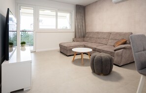 Ein modernes Wohnzimmer mit einem Ecksofa, einem kleinen runden Tisch, einem grauen Hocker und einem Flachbildfernseher auf einer weißen Kommode.