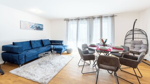 Een woonkamer met een blauwe bank, een eettafel met stoelen, een hangstoel en een schilderij aan de muur.
