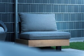 A modern outdoor seating area with a cushioned bench and a chair, set against a backdrop of a patterned wall and a grassy area.