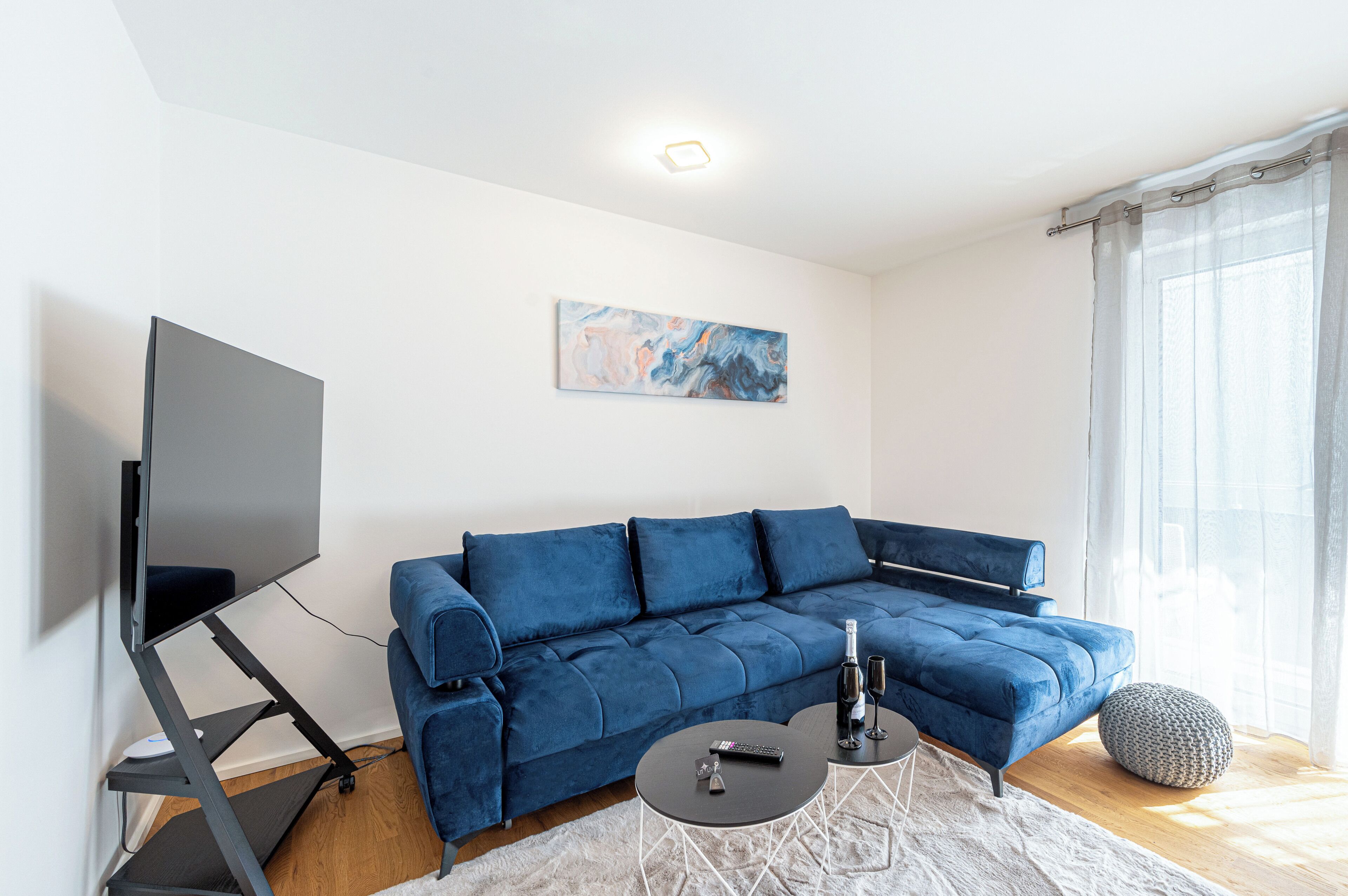 Un salon moderne avec un canapé d’angle bleu, un téléviseur à écran plat posé sur un meuble, une table basse et un tableau accroché au mur.
