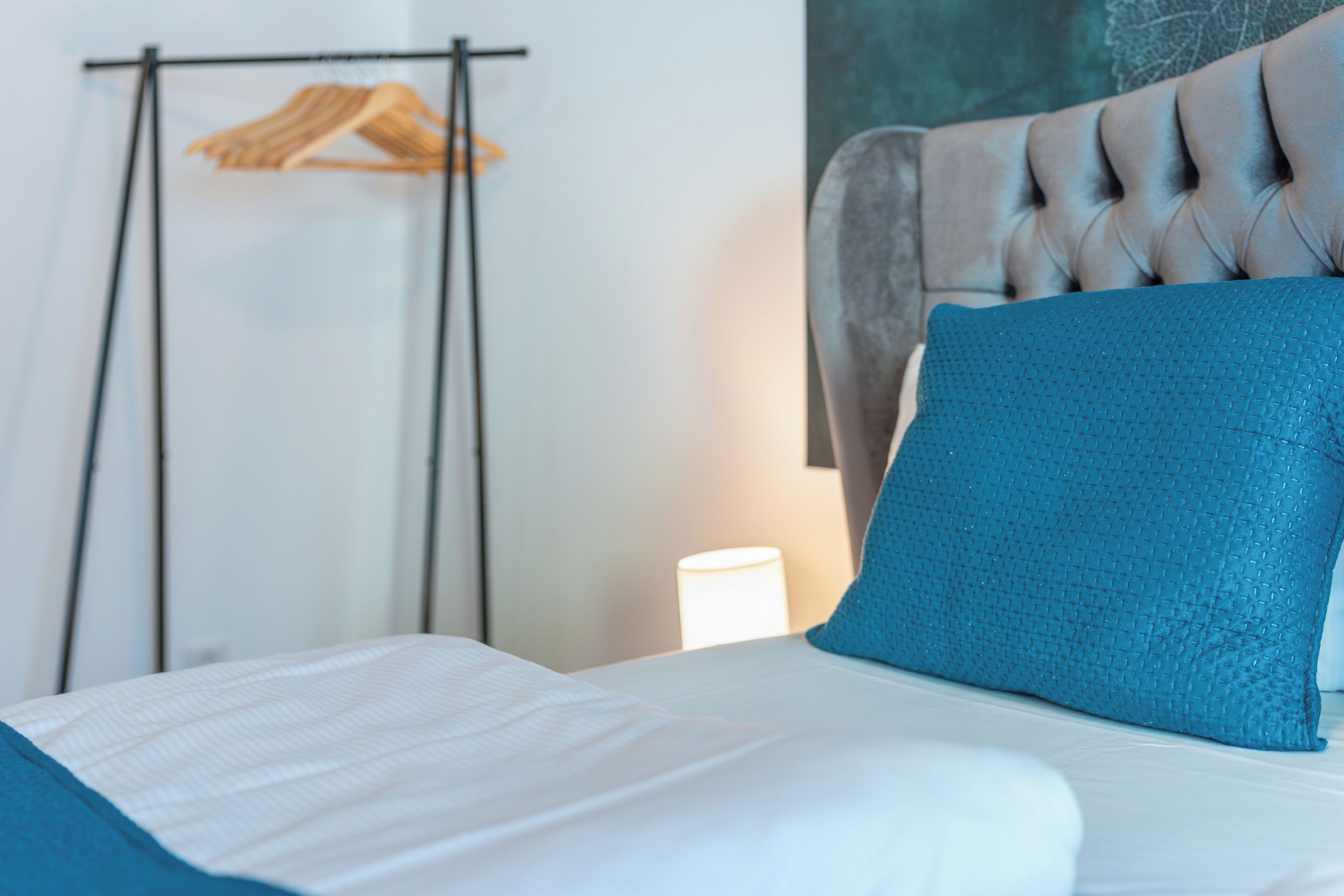 Een net opgemaakt bed met een blauw kussen, een kledingrek en een gestoffeerd hoofdeinde.