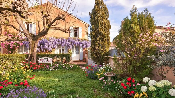 Ein Garten mit verschiedenen Blumen, einer Bank und einem Gebäude mit schrägem Dach.