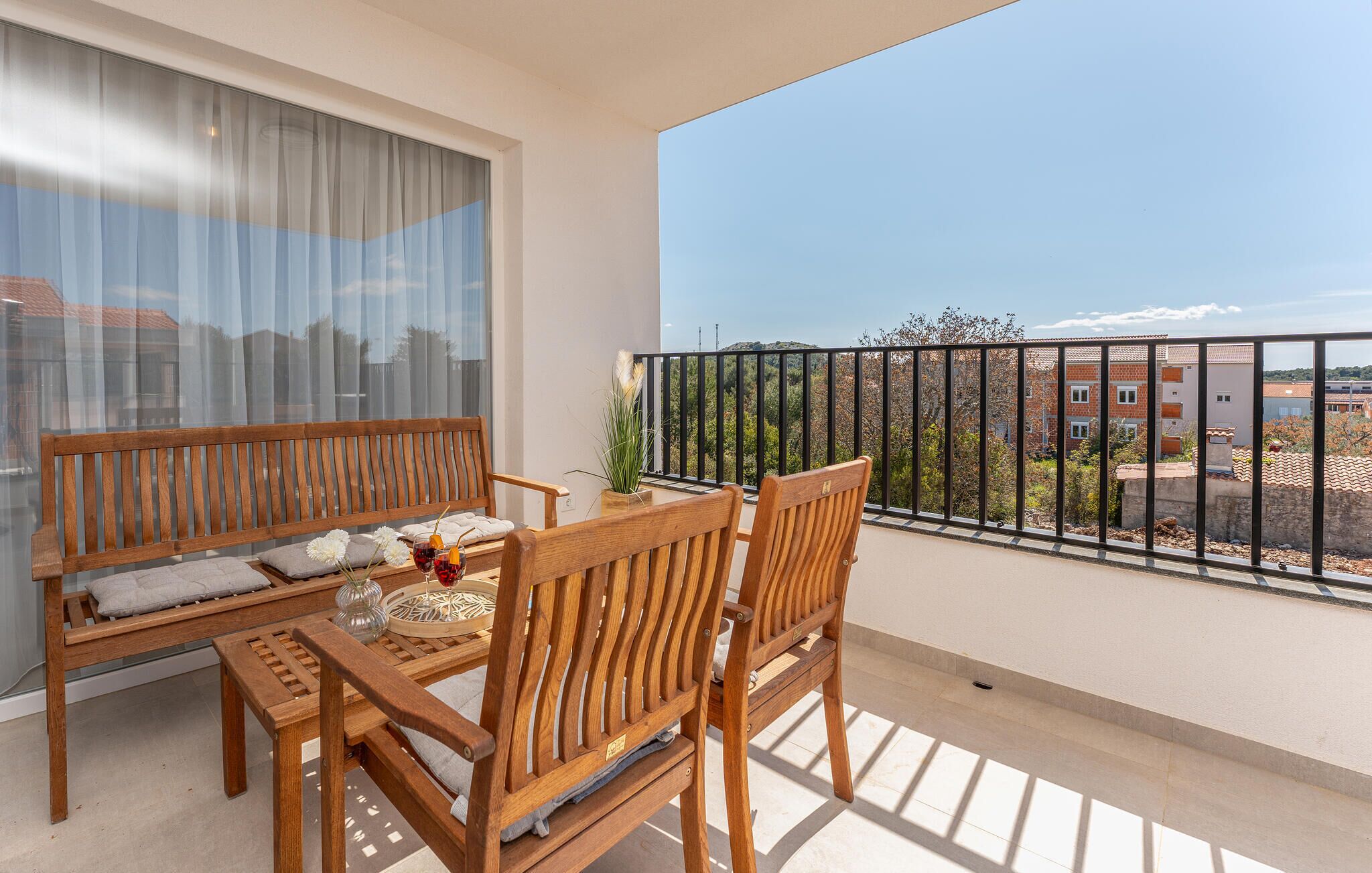 A balcony with a wooden bench and table, a potted plant, and a view of buildings and trees.
