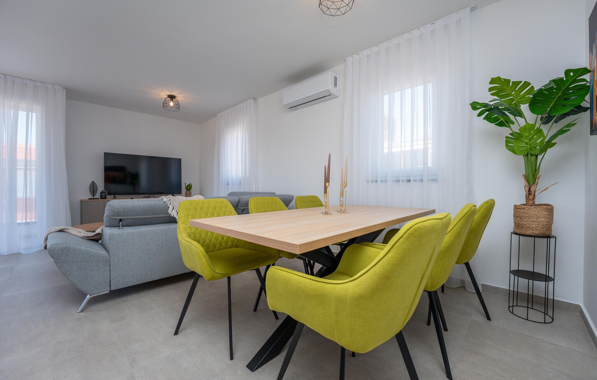 A modern living room with a dining area, grey sofa, yellow chairs, a TV, and a potted plant.