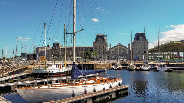Uma marina com barcos atracados, um edifício grande com uma torre do relógio e uma área coberta com assentos.