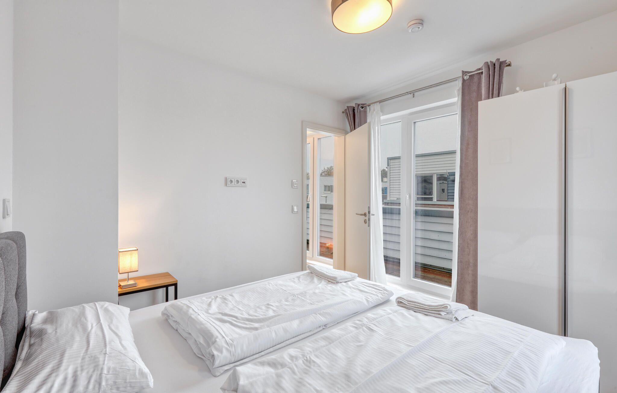 A hotel room with two beds, a bedside table with a lamp, a wardrobe, and a window with curtains.
