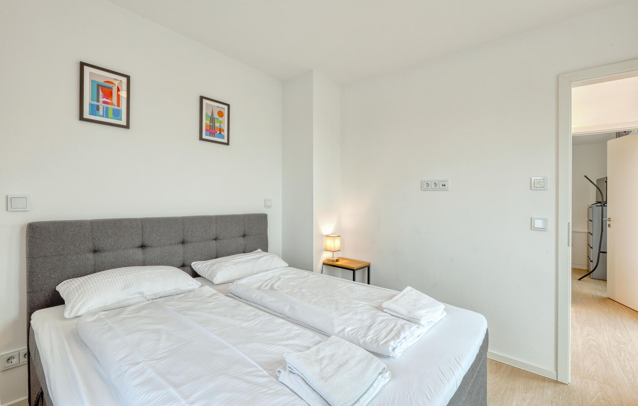 Una habitación de hotel con un cabecero tapizado en gris, dos camas individuales con sábanas blancas, una mesita de noche con lámpara y dos cuadros en la pared.