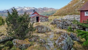 Une cabane en pierre avec un toit rouge, entourée d’un terrain rocailleux et d’une végétation clairsemée, avec une chaîne de montagnes en arrière-plan.