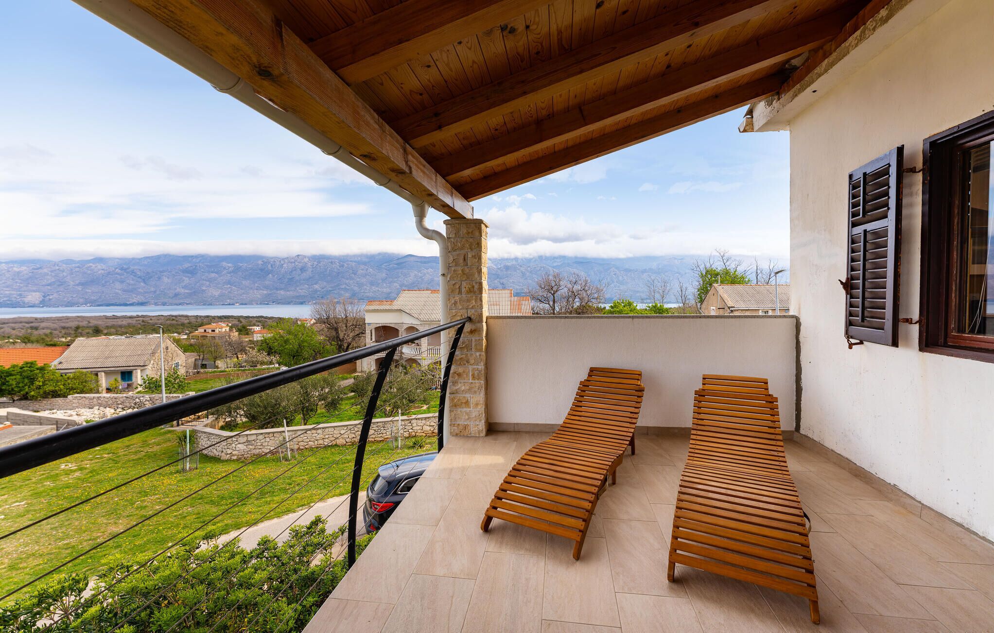 A balcony with two wooden benches, a car parked outside, and a view of a lake and mountains in the background.