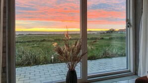 Blick durch ein Fenster auf einen Rasen mit Sonnenuntergang, mit Ziegelboden und einer Vase mit getrockneten Pflanzen.