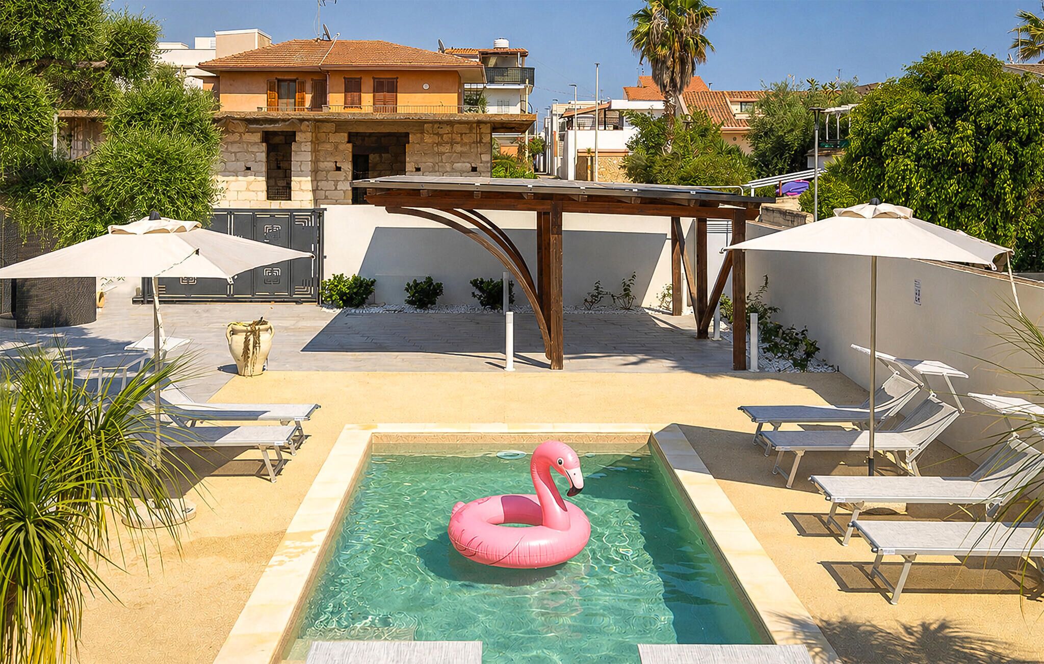 A swimming pool with a pink flamingo float, surrounded by sun loungers and umbrellas, with a stone building in the background.