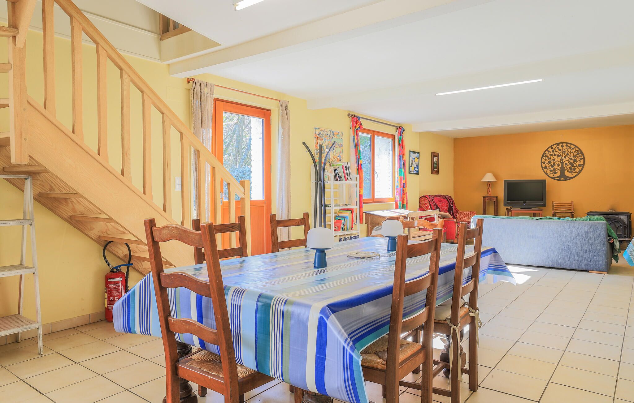 Una zona de comedor con una escalera de madera, una mesa con sillas, una estantería y un televisor al fondo.