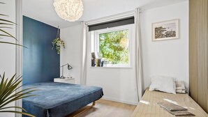 Et soveværelse med seng, sengebord, lampe, vindue med udsigt til grønne omgivelser og et billede i ramme på væggen.