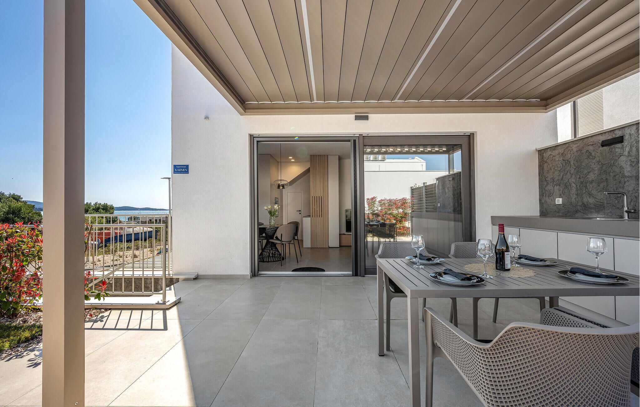 Een modern terras buiten, met een eettafel gedekt voor twee personen, een barbecue en uitzicht op zee.