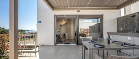 Een modern terras buiten, met een eettafel gedekt voor twee personen, een barbecue en uitzicht op zee.