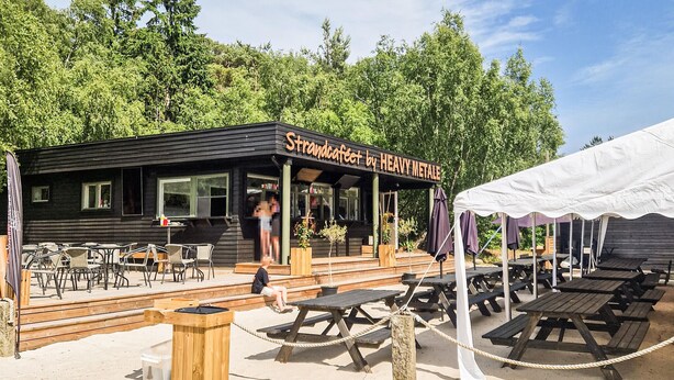 A wooden building with a sign reading "Strandcafeet by Heavy Metal" and outdoor seating with tables and chairs.