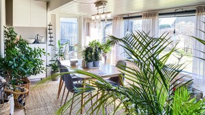 A dining area with a wooden table, chairs, potted plants, and a view of the outdoors through large windows.