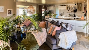A modern living room with a grey sofa, various cushions, and a large green plant. A kitchen with white cabinets and a stainless steel range hood is visible in the background.