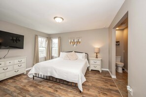 Een slaapkamer met een bed, een dressoir, een nachtkastje, een aan de muur bevestigde televisie en een raam met gordijnen.