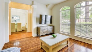 Un amplio salón con piso de madera, un televisor de pantalla plana montado en la pared y una zona de comedor con mesa y sillas.