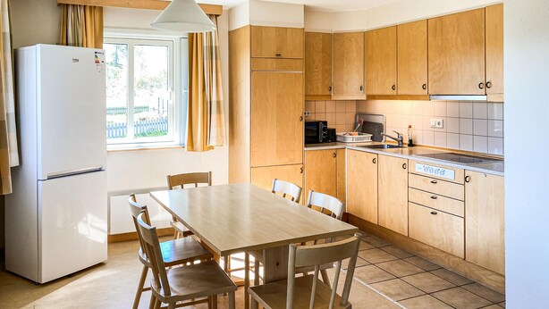 Une cuisine de taille réduite, avec des meubles de rangement en bois, un réfrigérateur blanc, une table à manger avec des chaises et une fenêtre agrémentée de rideaux.
