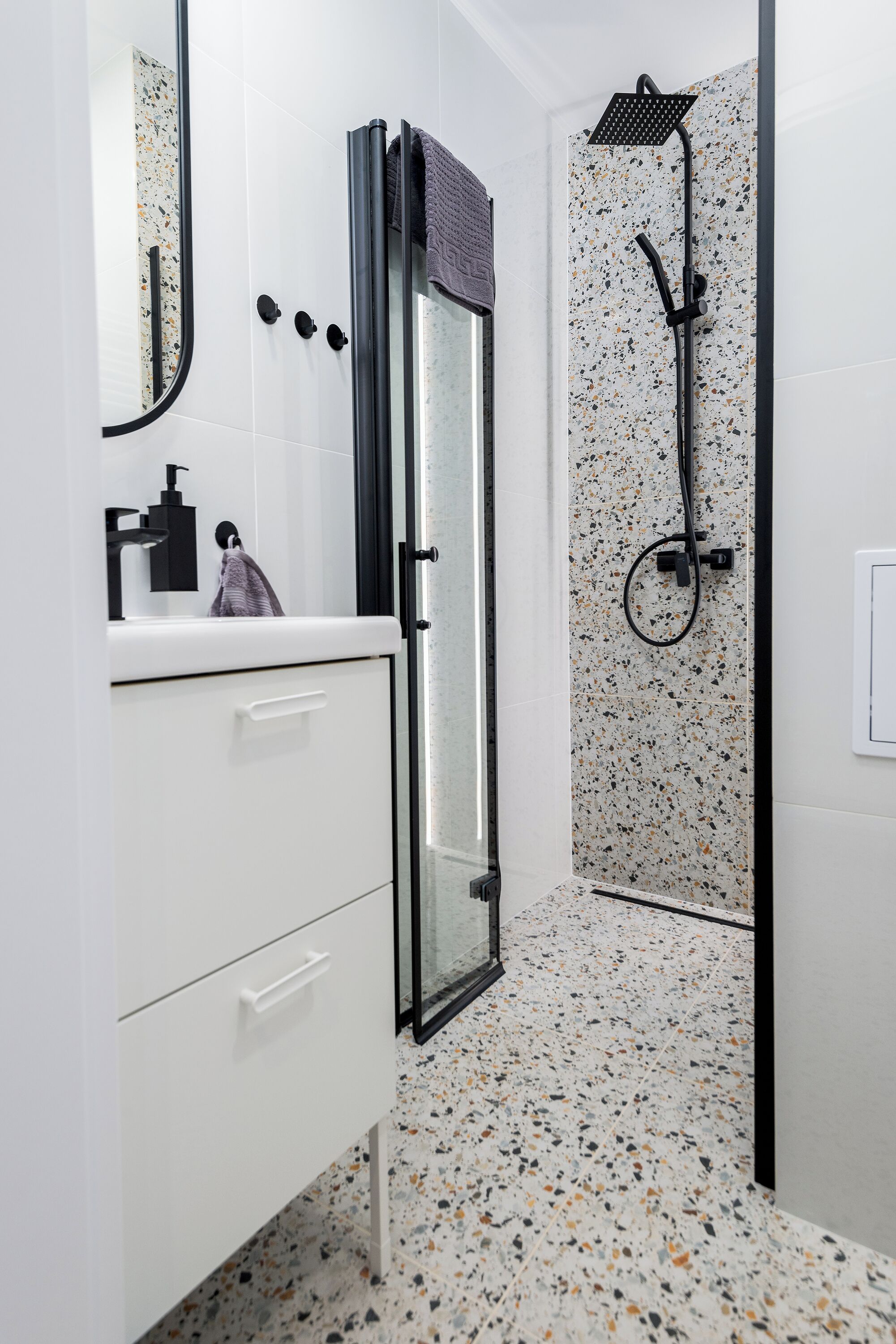 A modern bathroom with a glass shower enclosure, a white sink with a black faucet, and a speckled tile floor.